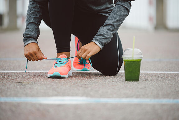 Eine Frau bindet sich ihren Turnschuh zu. Neben ihr steht ein gesunder Smoothie.